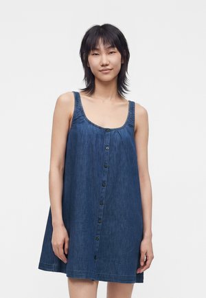 Kvinde står foran en ensfarvet baggrund iført en ærmeløs, løstsiddende blå denimkjole med knapper foran og kigger mod kameraet.