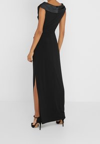 Dlouhé černé šaty se saténovým drapovaným límcem, přiléhavým střihem, bočním rozparkem a černými vysokými podpatky. Hladká textilie.