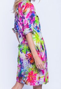 Bunt gemustertes Blumenkleid mit kurzen Ärmeln, das lebendige Rosa-, Grün- und Blautöne auf leichtem Stoff zeigt. Seitenschlitze sorgen für Bewegung.