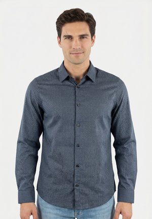 Uomo che indossa una camicia blu scuro a maniche lunghe con fantasia e jeans azzurri chiari, in piedi davanti a uno sfondo bianco semplice.