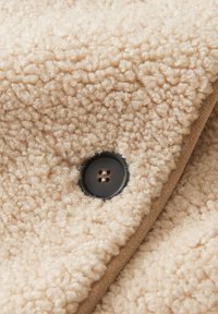 Beiger Fleece-Stoff mit einer strukturierten, flauschigen Oberfläche; verfügt über einen schwarzen Knopf, der zum Verschließen in das Material genäht ist.
