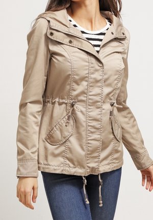 Vrouw draagt een beige capuchonvest met voorzakken en een taillekoord, gecombineerd met donkerblauwe spijkerbroek en een zwart-wit gestreept shirt.