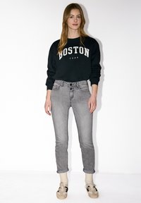 Femme debout contre un mur blanc, portant un sweat-shirt noir "Boston", un jean gris retroussé, des chaussettes crème et des baskets beiges.
