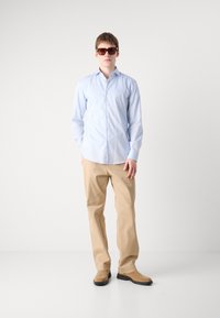 Light blue button-up shirt, beige pants, and brown suede shoes. Model wearing dark sunglasses, standing against a plain background.