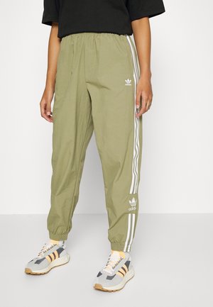 Personne portant un pantalon de survêtement Adidas vert olive avec des bandes blanches et des logos, un t-shirt noir, et des baskets grises et beiges avec des touches jaunes.