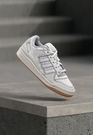 Baskets blanches Adidas avec semelle en gomme et trois bandes, suspendues au-dessus d'un rebord en béton avec des escaliers flous en arrière-plan.