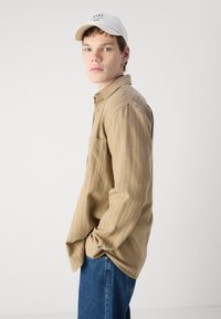 Cămașă bej cu mânecă lungă și dungi verticale, dotată cu un buzunar pe piept, purtată cu blugi albaștri și o șapcă albă cu un logo.