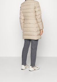 Beige pufferjas met horizontale stitching, lange mouwen en een recht model. Gecombineerd met grijze jeans en witte sneakers. Geen zichtbare details.