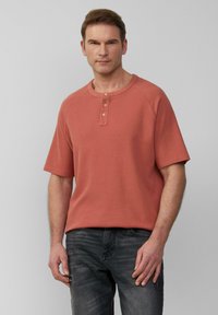 Koszulka henley z krótkim rękawem w kolorze koralowym z trzema drewnianymi guzikami, okrągłym dekoltem i teksturowanym materiałem; zestawiona z ciemnymi dżinsami.