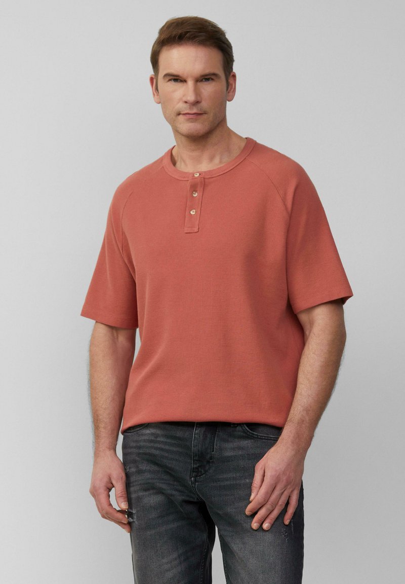 Koszulka henley z krótkim rękawem w kolorze koralowym z trzema drewnianymi guzikami, okrągłym dekoltem i teksturowanym materiałem; zestawiona z ciemnymi dżinsami.