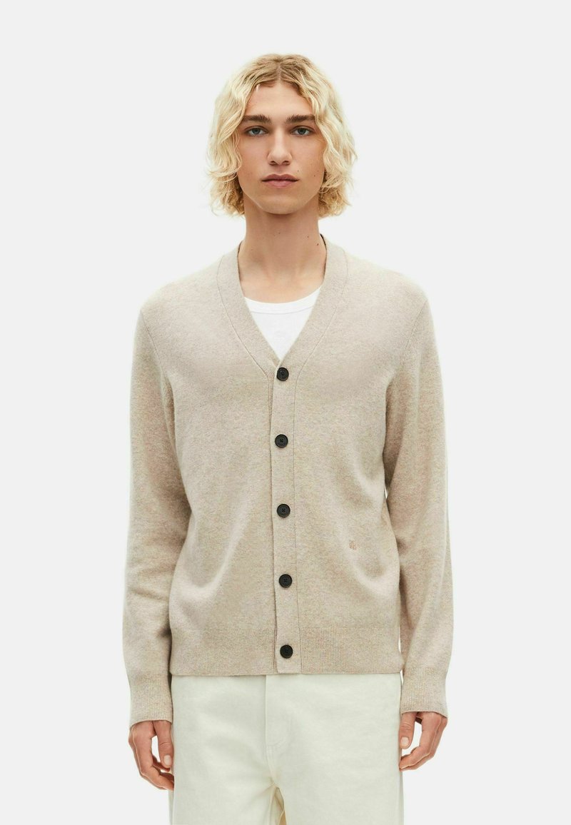 Beige cardigan van zacht materiaal, met een V-hals, zwarte knopen en lange mouwen. Draagt over een witte shirt, met subtiele textuur.