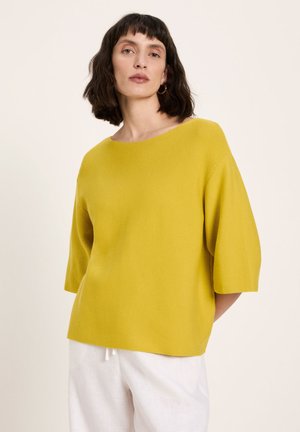 Woman wearing a mustard yellow boat neck sweater with elbow-length sleeves and white pants against a plain background.