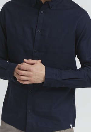 Mężczyzna w długim, granatowym koszuli zapinanej na guziki, ze splecionymi rękami na wysokości talii, stojący na jednolitym, jasnym tle.