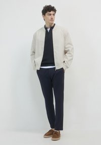 Giovane uomo che indossa una giacca bomber beige, un maglione blu navy e pantaloni, con scarpe in camoscio marroni, in piedi con le mani in tasca, mentre guarda a sinistra.