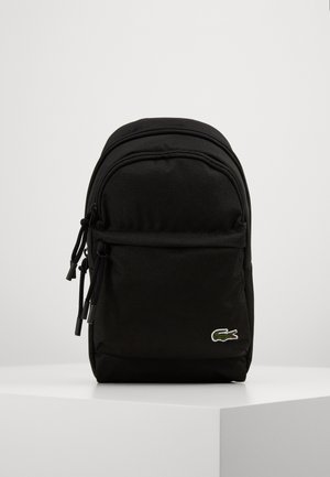 Sac à dos compact noir avec poche avant zippée et logo crocodile vert Lacoste en bas à droite, posé sur une surface blanche.