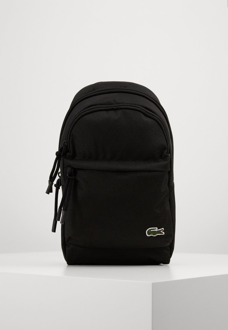 Sac à dos compact noir avec poche avant zippée et logo crocodile vert Lacoste en bas à droite, posé sur une surface blanche.