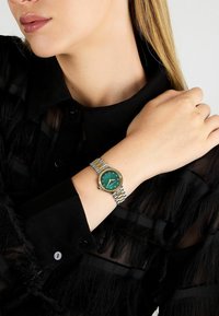 Montre-bracelet bicolore en or et argent avec un cadran vert, des index dorés et un boîtier rond, portée sur un poignet avec une chemise noire.