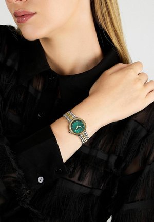 Orologio bracciale bicolore oro e argento con quadrante verde, indici delle ore dorati e cassa rotonda, indossato su un polso con una camicia nera.