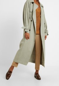 Langer, hellgrüner Leinen-Trenchcoat mit doppelreihigen Knöpfen und umgeschlagenen Ärmeln, kombiniert mit einem braunen Strickoberteil und beigen Hosen, dazu braune Mokassins.
