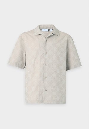 Homme portant une chemise beige à manches courtes à motifs et un pantalon ample assorti avec des baskets noires et blanches, debout devant un fond blanc.
