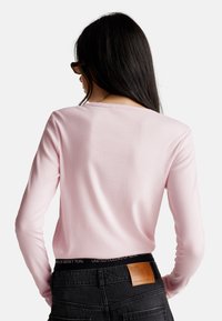 Kobieta z długimi ciemnymi włosami, ubrana w różową bluzkę z długim rękawem, czarne dżinsy z skórzaną łatką i okulary przeciwsłoneczne w kolorze żółwia, stoi tyłem.