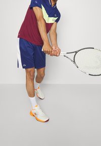Herrernas Lacoste sportkläder med en vinröd och blå kortärmad skjorta, blå shorts och vit-orange skoset, håller en tennisracket.