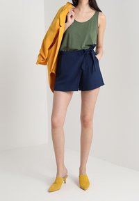 Woman wearing green tank top, navy blue tied shorts, yellow heels, holding a mustard blazer over one shoulder against a white background.