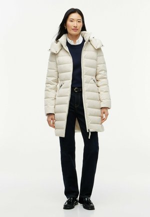 Kvinde stående iført en beige lang dunjakke, sorte bukser, sorte sko, en marineblå sweater og en hvid skjorte med krave.