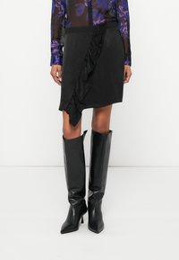 Fekete szoknya fodros oldaldísszel, sima anyagból. Térdig érő fekete bőr csizmával párosítva, hegyes orrú és sarkú.