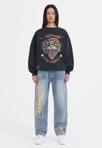 Zwarte sweatshirt met een tijgerafbeelding en tekst, gecombineerd met vervaagde blauwe wijde jeans met gele "Ed Hardy" letters en witte sneakers.