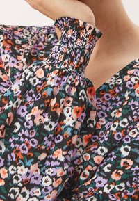 Blusi kukkaprintillä, jossa on musta tausta, puffatut hihat, joissa on rypytetyt hihansuut, ja eloisa monivärinen kuviointi purppuran, oranssin ja valkoisen sävyissä.