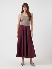 Donna che indossa un top senza maniche beige, una gonna midi borgogna e scarpe con tacco basso color taupe, in piedi su uno sfondo bianco.