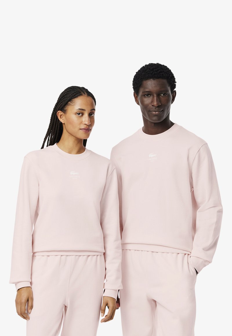 Felpe rosa chiaro con maniche lunghe presentano un collo rotondo e un logo frontale. Il tessuto sembra morbido e sono abbinate a pantaloni coordinati.