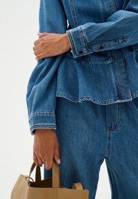 Blauwe spijkerjas met gerimpelde zoom, gedetailleerde stiksels en knopen. Persoon houdt een tan leren handtas vast. Close-up van de arm en de outfit.