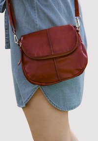 Rode leren crossbody tas met een klepsluiting, één ritsvak en een verstelbare schouderriem, tentoongesteld tegen een lichtblauwe denim kledingstuk.