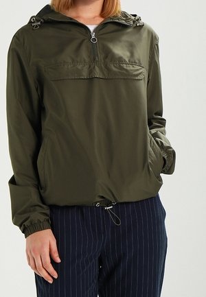 Personne portant un coupe-vent à capuche vert olive avec fermeture éclair avant et poches, associé à un pantalon bleu marine à fines rayures.