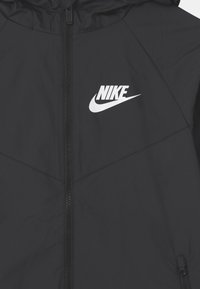 Musta Nike-takki, jossa on vetoketju edessä ja valkoinen logo vasemmassa rinnassa. Valmistettu kevyestä materiaalista, jossa sileä pinta.