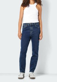 Tmavě modré džíny s rovnými nohavicemi, které mají pět kapes a knoflíkový zip, ve spojení s bílým topem bez rukáv ů.