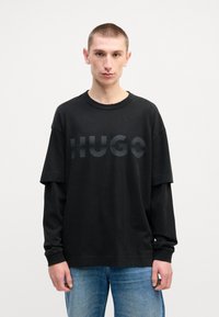 Juodas oversized marškinėliai su toniniu "HUGO" logotipu. Pagaminti iš minkštos medžiagos, turi nuleistus pečius ir laisvą siluetą. Derinami su mėlynomis džinsais.