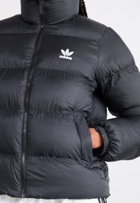 Zwarte pufferjack met hoge kraag, ritssluiting, elastische manchetten en zijzakken. Bevat het witte Adidas-logo op de borst. Gladde textuur.