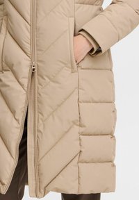 Parka beige trapuntata con un motivo a zigzag diagonale, dotata di una chiusura frontale con zip e tasche laterali. Tessuto liscio con una consistenza morbida.