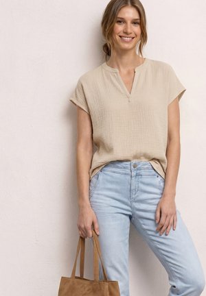 Uśmiechnięta kobieta w beżowej bluzce z krótkim rękawem i jasnoniebieskich dżinsach trzyma beżową torbę, stojąc na tle jednolitej jasnej ściany.