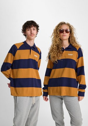 Två personer klädda i mörkblå och senapsfärgade randiga långärmade polotröjor och ljusgrå sweatpants, stående mot en vit bakgrund.