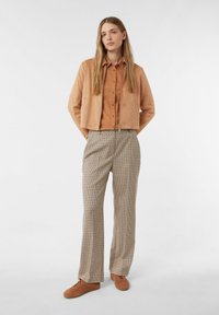 Donna in piedi con le mani dietro la schiena, indossa una giacca di camoscio beige, una camicia rossa con bottoni, pantaloni a quadri e sneaker marroni su uno sfondo neutro.