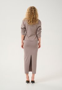 Das Produkt zeichnet sich durch ein eng anliegendes Langarmoberteil und einen Bleistiftrock aus weichem Taupe-Stoff mit einem Schlitz auf der Rückseite aus, kombiniert mit eleganten schwarzen High Heels.