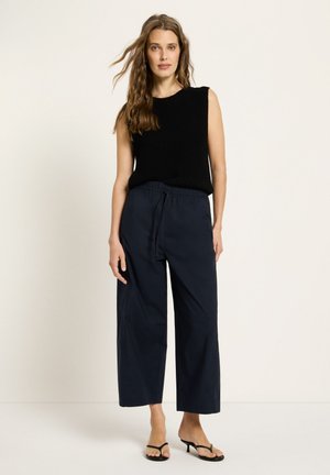 Woman with long hair wears sleeveless black knit top, navy wide-leg cropped pants, and black strappy heeled sandals against plain background.