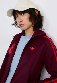 Burgundská zipová bunda s červenými pruhy, s logy Adidas a modrým tričkem pod ní, doplněná bílou čepicí. Hladká textilie.