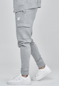 Graue Baumwoll-Jogginghosen mit elastischen Bündchen, ausgestattet mit einer Seitentasche und einem weißen Logo. Kombiniert mit weißen Sportschuhen.