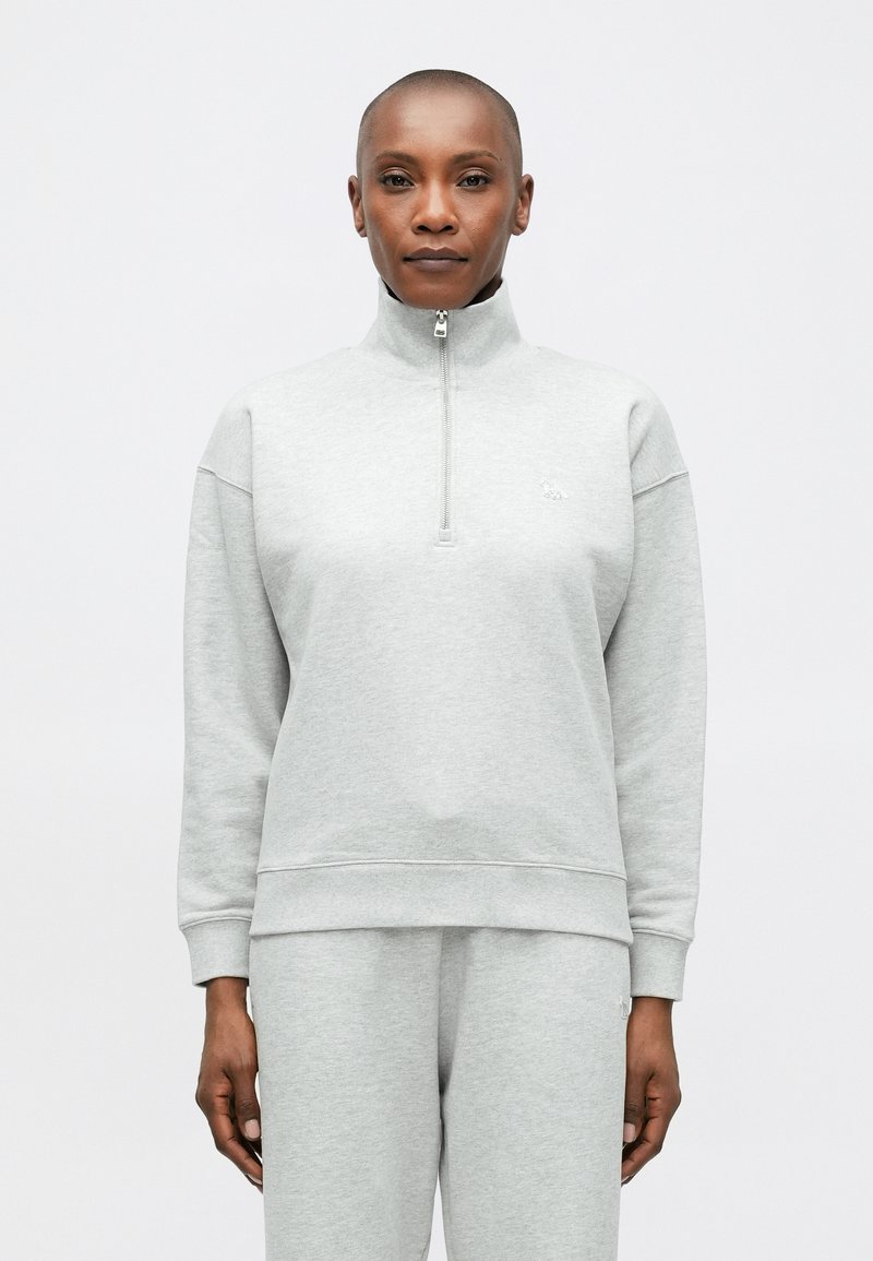 Svetlo šedá mikina na zips s vysokým golierom, ribovanými manžetami a lemom. Vyrobená z mäkkej tkaniny, s drobným vyšitým logom na hrudi.