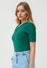 Model in een aansluitend groen topje met een V-hals en korte mouwen, gecombineerd met lichtblauwe high-waisted shorts, tegen een effen achtergrond.
